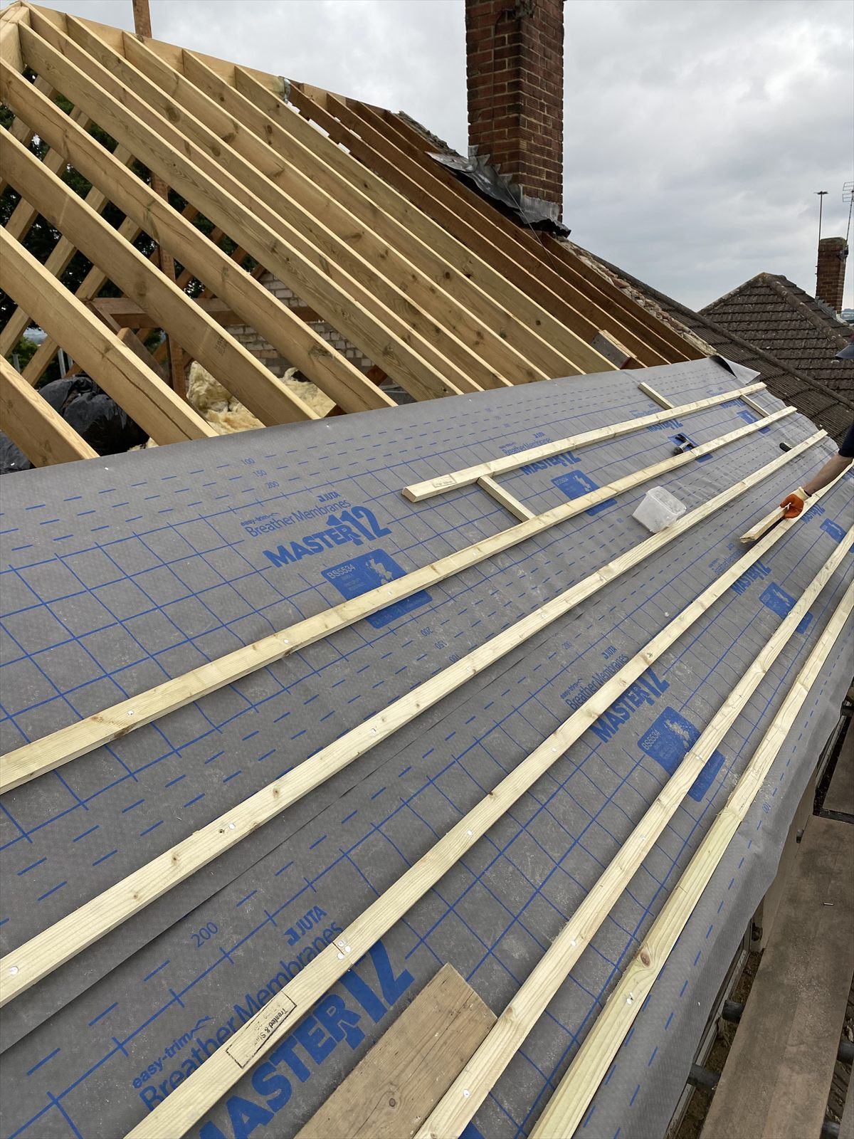 Roofer fitting breather membrane and battens to a newly framed roof, with a chimney stack behind